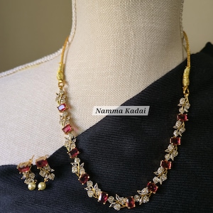 May include: A gold-toned necklace and earring set featuring red rectangular stones and small clear stones. The necklace and earrings have a floral design. The text "Namma Kadai" is visible on the image.