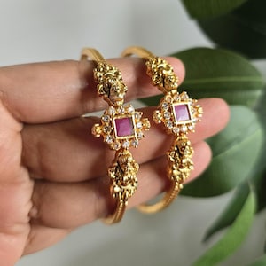 May include: Two gold-colored bangles with intricate detailing. Each bangle features a central square ruby-colored stone surrounded by small clear stones. The design includes ornate figures and textured bands.