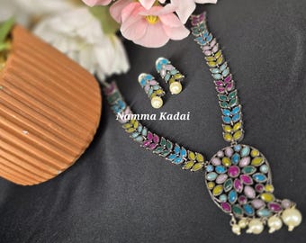 Puede incluir: Un collar de plata con un colgante colorido y pendientes a juego. El colgante tiene forma de lágrima y está decorado con piedras de colores. El collar es de plata y tiene una cadena con diseño de hojas. Los pendientes también son de plata y tienen un diseño de hojas. El texto "Namma Kadai" es visible en la imagen.