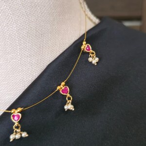 Puede incluir: Delicado collar de oro con tres colgantes en forma de coraz&oacute;n con centros de color rojo rub&iacute; y peque&ntilde;os grupos de perlas. El collar se muestra sobre un fondo de tela negra, resaltando el dise&ntilde;o intrincado y el contraste de color.