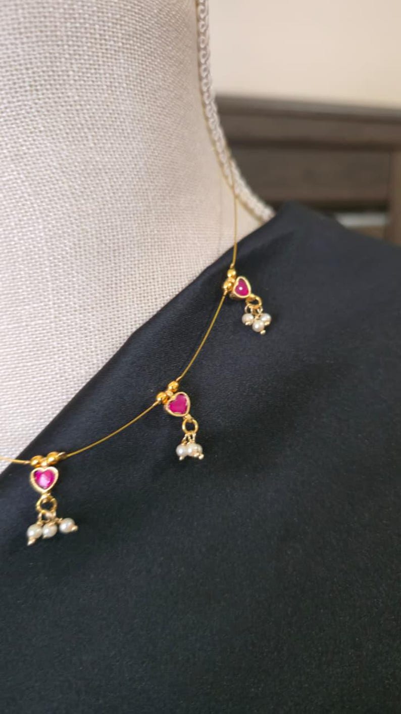 Puede incluir: Delicado collar de oro con tres colgantes en forma de coraz&oacute;n con centros de color rojo rub&iacute; y peque&ntilde;os grupos de perlas. El collar se muestra sobre un fondo de tela negra, resaltando el dise&ntilde;o intrincado y el contraste de color.