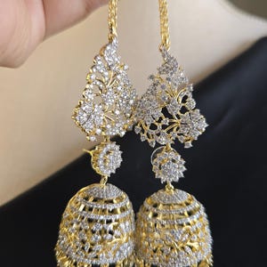 May include: A pair of gold-toned earrings with intricate floral and bell-shaped designs. The earrings feature sparkling clear stones and hang from delicate chains. The design is ornate and detailed, with a focus on elegance and sparkle.