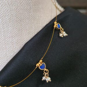 Puede incluir: Delicado collar dorado con colgantes azules en forma de coraz&oacute;n y peque&ntilde;os detalles de perlas blancas. El collar se muestra sobre un fondo negro, resaltando el dise&ntilde;o y el color de la joya.