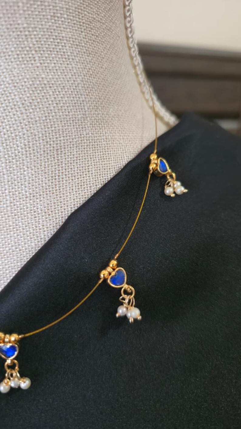 Puede incluir: Delicado collar dorado con colgantes azules en forma de coraz&oacute;n y peque&ntilde;os detalles de perlas blancas. El collar se muestra sobre un fondo negro, resaltando el dise&ntilde;o y el color de la joya.