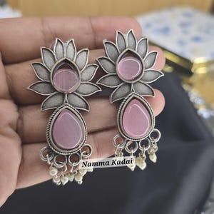 May include: A pair of silver earrings with a pink flower design and a pink teardrop-shaped stone hanging from each. The earrings have a silver chain with small white beads hanging from the bottom.