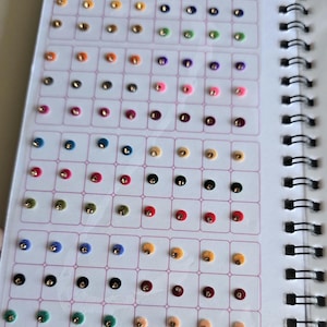 May include: A spiral-bound booklet containing rows of adhesive sticker kumkum dots in various colors, including red, blue, yellow, green, and black. Each small, circular sticker features a central metallic accent. The text A QUALITY PRODUCT and STICKER KUMKUM is printed at the top of the page.