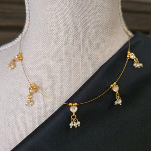 Puede incluir: Delicado collar dorado con colgantes en forma de coraz&oacute;n y peque&ntilde;os detalles de perlas. El collar se exhibe sobre un maniqu&iacute;, sobre un fondo de tela negra. El dise&ntilde;o es sencillo y elegante.