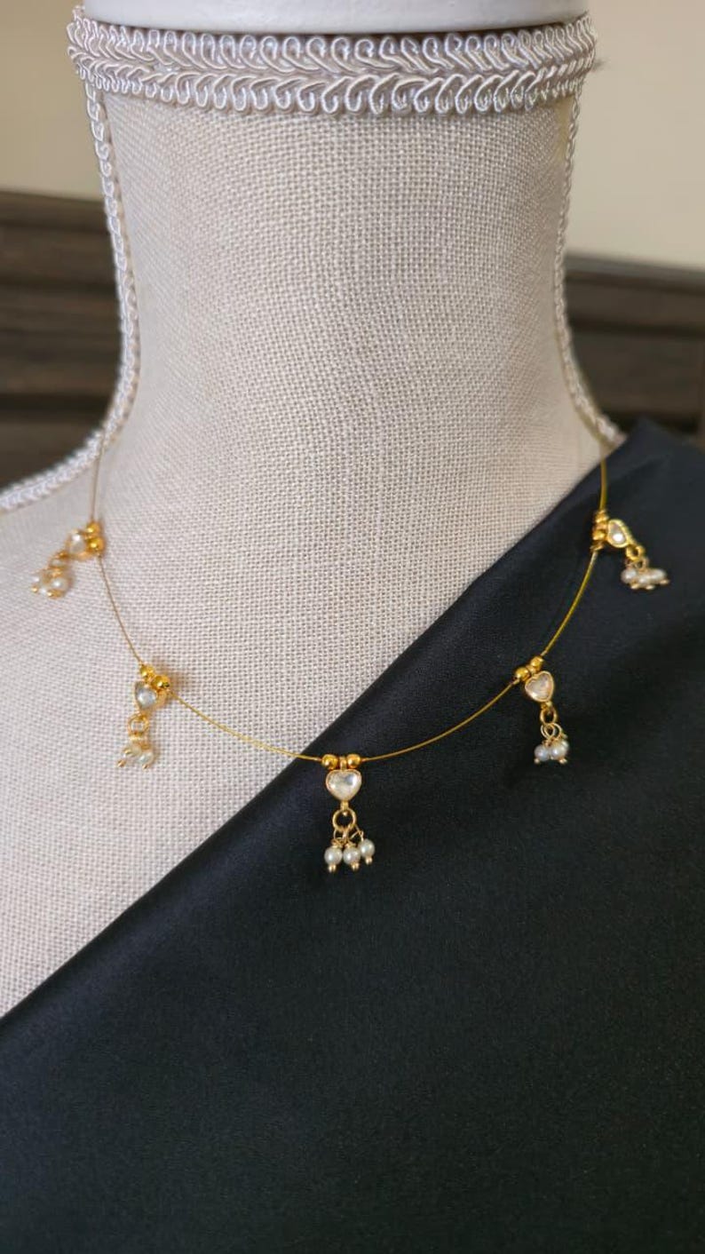 Puede incluir: Delicado collar dorado con colgantes en forma de coraz&oacute;n y peque&ntilde;os detalles de perlas. El collar se exhibe sobre un maniqu&iacute;, sobre un fondo de tela negra. El dise&ntilde;o es sencillo y elegante.