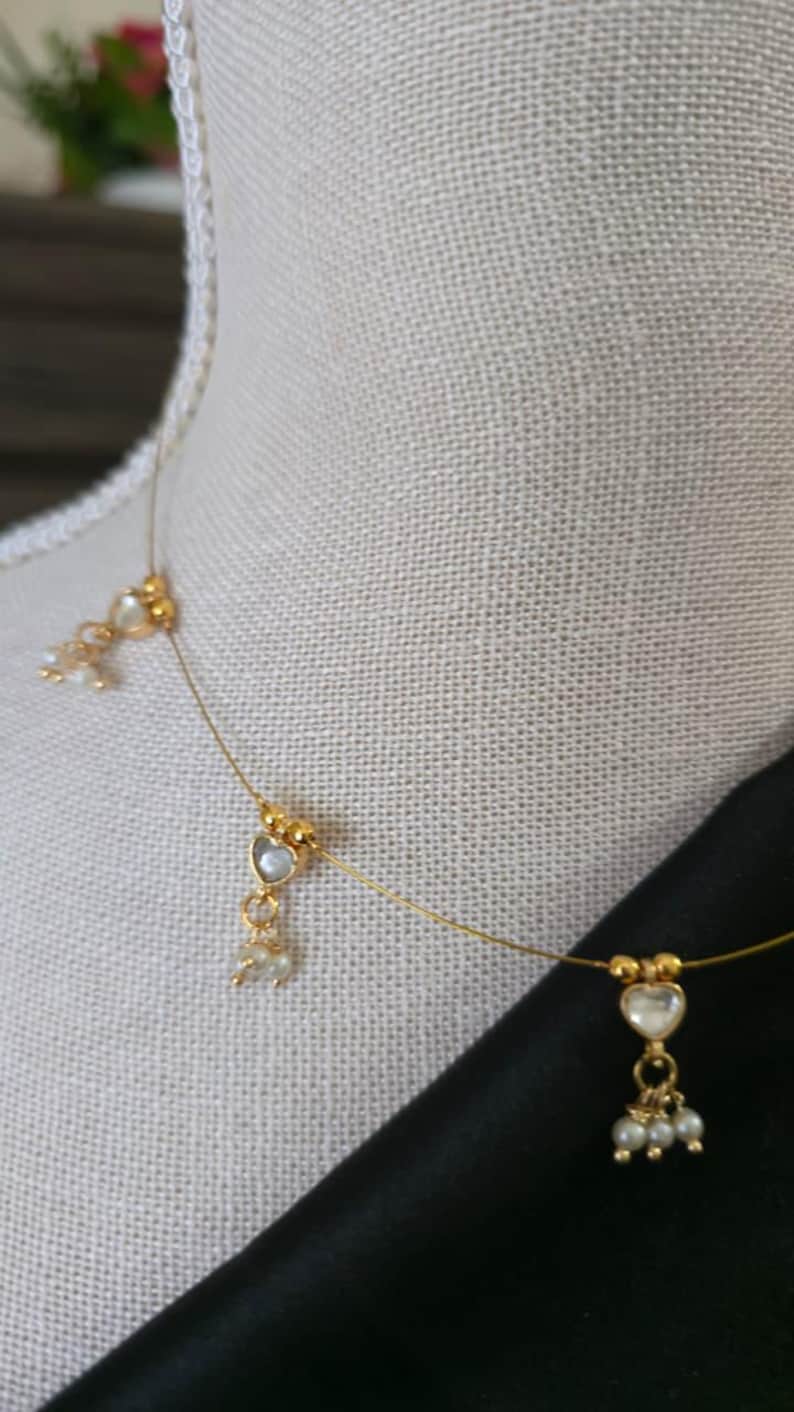 Puede incluir: Delicado collar dorado con colgantes en forma de coraz&oacute;n y peque&ntilde;os detalles de perlas. El collar se exhibe sobre un maniqu&iacute; con un fondo de tela de color neutro. El dise&ntilde;o es sencillo y elegante, adecuado para diversas ocasiones.