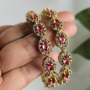May include: Two gold bangles with oval, ruby-colored gemstones and small, clear accent stones. The bangles are held in a hand, with a soft background. The jewelry has a vintage or antique aesthetic.