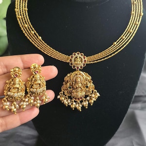 May include: A gold-toned necklace featuring five strands and a central pendant depicting a seated deity, accented with small red and green stones and white beads. Matching drop earrings feature the same deity motif and bead accents.