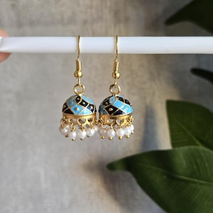 May include: A pair of gold-tone jhumka earrings featuring a bell-shaped design with intricate black and light blue enamel patterns. The base of each earring is adorned with a scalloped gold edge and a row of small, dangling white faux pearls.