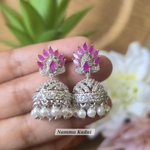 May include: A pair of silver-toned jhumka earrings with pink gemstone accents and dangling white pearls. The earrings have a detailed, ornate design. The text "Namma Kadai" is visible on the image.