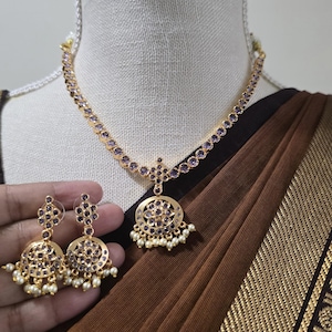May include: A gold-toned necklace and matching earring set featuring circular pendants inlaid with small purple stones and accented with dangling white pearls. The necklace chain is also adorned with a series of small, round purple stones.