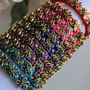 May include: A stack of multi-colored bangles wrapped in silk thread, featuring gold-toned metal floral overlays with embedded colorful crystals. The bangles are arranged in a gradient of red, pink, purple, and blue, showcasing intricate petal designs.
