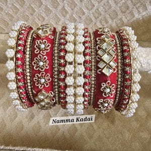 May include: A collection of red and gold bangles with intricate floral and geometric designs. The bangles are adorned with pearls and gemstones, and the text "Namma Kadai" is visible.