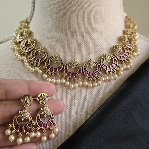 May include: Gold-toned necklace and matching earrings set. The necklace features a series of ornate, circular pendants with a deep pink center and pearl accents. The earrings mirror the necklace design, with a similar circular shape, pink stones, and dangling pearls.