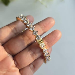 Puede incluir: Una pulsera de tenis en tono dorado que presenta una hilera continua de piedras redondas, transparentes y facetadas, engastadas en garras met&aacute;licas individuales. La pulsera incluye un mecanismo de cierre de bisagra visible en el centro.