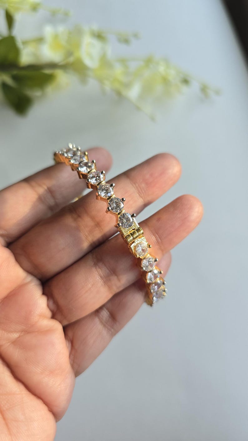 Puede incluir: Una pulsera de tenis en tono dorado que presenta una hilera continua de piedras redondas, transparentes y facetadas, engastadas en garras met&aacute;licas individuales. La pulsera incluye un mecanismo de cierre de bisagra visible en el centro.