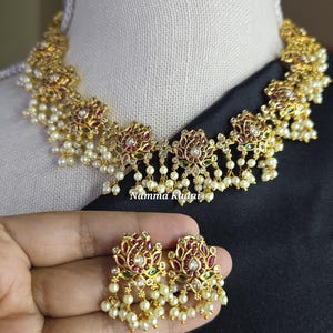 May include: Gold-toned necklace and earring set with floral and pearl accents. The necklace and earrings feature red, green, and clear gemstones. The earrings are held in a hand. The text "Namma Kudai" is visible on the necklace.