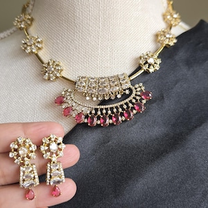 May include: Gold-toned necklace and earring set featuring clear and ruby-red gemstones. The necklace has a floral design with a pendant of rectangular and teardrop-shaped stones. The earrings mirror the pendant's design.