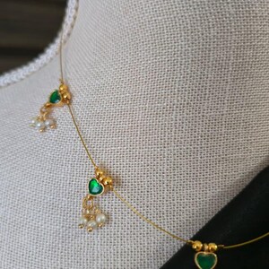 Puede incluir: Delicado collar de oro con piedras verdes en forma de coraz&oacute;n y peque&ntilde;as perlas blancas. El collar se exhibe en un maniqu&iacute;, mostrando su elegante dise&ntilde;o. El collar tiene un estilo minimalista, perfecto para el uso diario o ocasiones especiales.