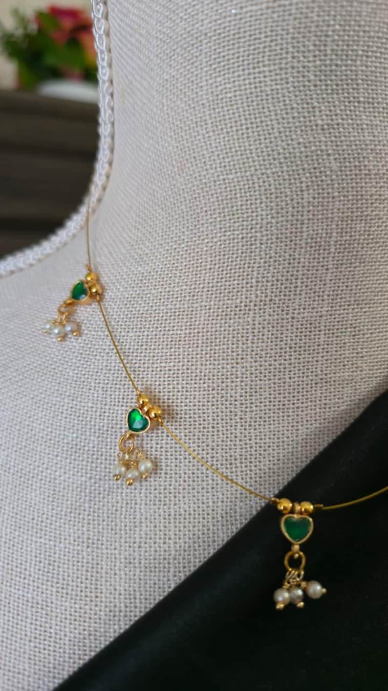 Puede incluir: Delicado collar de oro con piedras verdes en forma de coraz&oacute;n y peque&ntilde;as perlas blancas. El collar se exhibe en un maniqu&iacute;, mostrando su elegante dise&ntilde;o. El collar tiene un estilo minimalista, perfecto para el uso diario o ocasiones especiales.