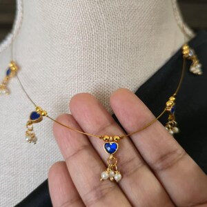 Puede incluir: Delicado collar dorado con colgantes azules en forma de coraz&oacute;n y peque&ntilde;os detalles de perlas blancas. El collar se exhibe en un maniqu&iacute;, mostrando su dise&ntilde;o. Una joya delicada y elegante.