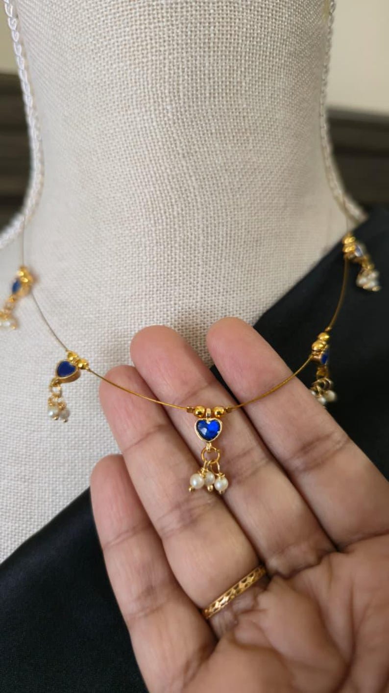 Puede incluir: Delicado collar dorado con colgantes azules en forma de coraz&oacute;n y peque&ntilde;os detalles de perlas blancas. El collar se exhibe en un maniqu&iacute;, mostrando su dise&ntilde;o. Una joya delicada y elegante.