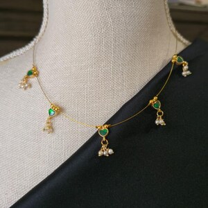 Puede incluir: Delicado collar de oro con colgantes verdes en forma de coraz&oacute;n y peque&ntilde;os detalles de perlas. El collar se exhibe sobre un maniqu&iacute;, sobre un fondo de tela negra. Una pieza de joyer&iacute;a sencilla y elegante.
