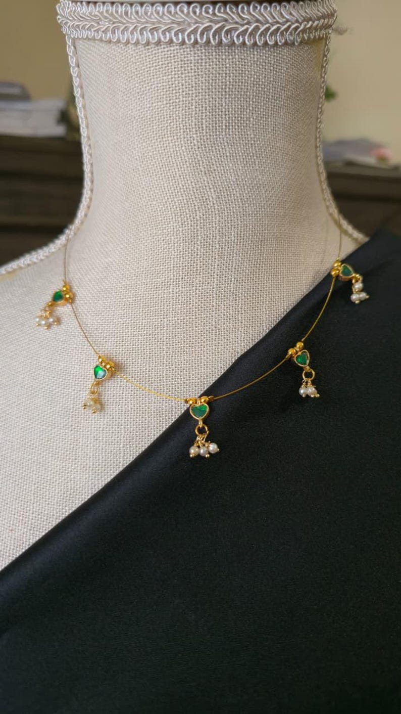 Puede incluir: Delicado collar de oro con colgantes verdes en forma de coraz&oacute;n y peque&ntilde;os detalles de perlas. El collar se exhibe sobre un maniqu&iacute;, sobre un fondo de tela negra. Una pieza de joyer&iacute;a sencilla y elegante.