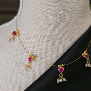 Puede incluir: Delicado collar de oro con colgantes de rub&iacute;es en forma de coraz&oacute;n y peque&ntilde;os grupos de perlas blancas. El collar se exhibe sobre un maniqu&iacute;, con una tela negra de fondo. El collar es una joya.