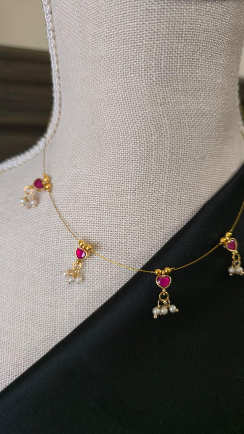 Puede incluir: Delicado collar de oro con colgantes de rub&iacute;es en forma de coraz&oacute;n y peque&ntilde;os grupos de perlas blancas. El collar se exhibe sobre un maniqu&iacute;, con una tela negra de fondo. El collar es una joya.