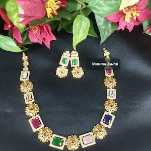 May include: A gold necklace with a row of colorful square stones and floral accents. The necklace is paired with matching earrings with green square stones and floral accents. The text "Namma Kadai" is visible in the image.