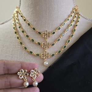 May include: A gold-toned necklace with three strands of green oval-shaped stones and flower pendants. Matching flower earrings with pearl drops are also visible. The jewelry is displayed on a mannequin.