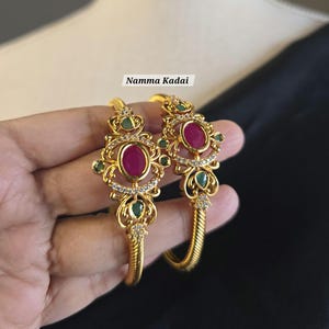 May include: Two gold-colored bangles with intricate designs. Each bangle features a large, oval, ruby-colored stone at the center, surrounded by smaller green stones and clear crystals. The text "Namma Kadai" is visible.