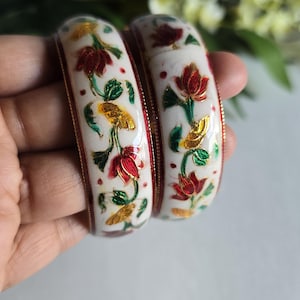 May include: Two white bangles featuring a hand-painted floral design with red, green, and gold metallic accents. The bracelets have a smooth, rounded surface and are finished with thin, dark red metallic trim along the edges of each piece.