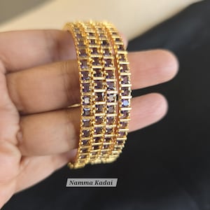 May include: Gold-toned bangles with square, purple gemstones. The bangles are stacked, showcasing a repeating pattern of the gemstones. The text "Namma Kadai" is visible on the image. These bangles are a type of jewelry.