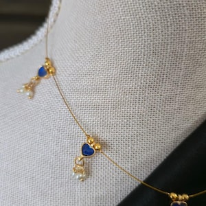 Puede incluir: Delicado collar de oro con colgantes azules en forma de coraz&oacute;n y peque&ntilde;os detalles de perlas blancas. El collar se exhibe en un maniqu&iacute;, mostrando su elegante dise&ntilde;o. Una joya elegante.