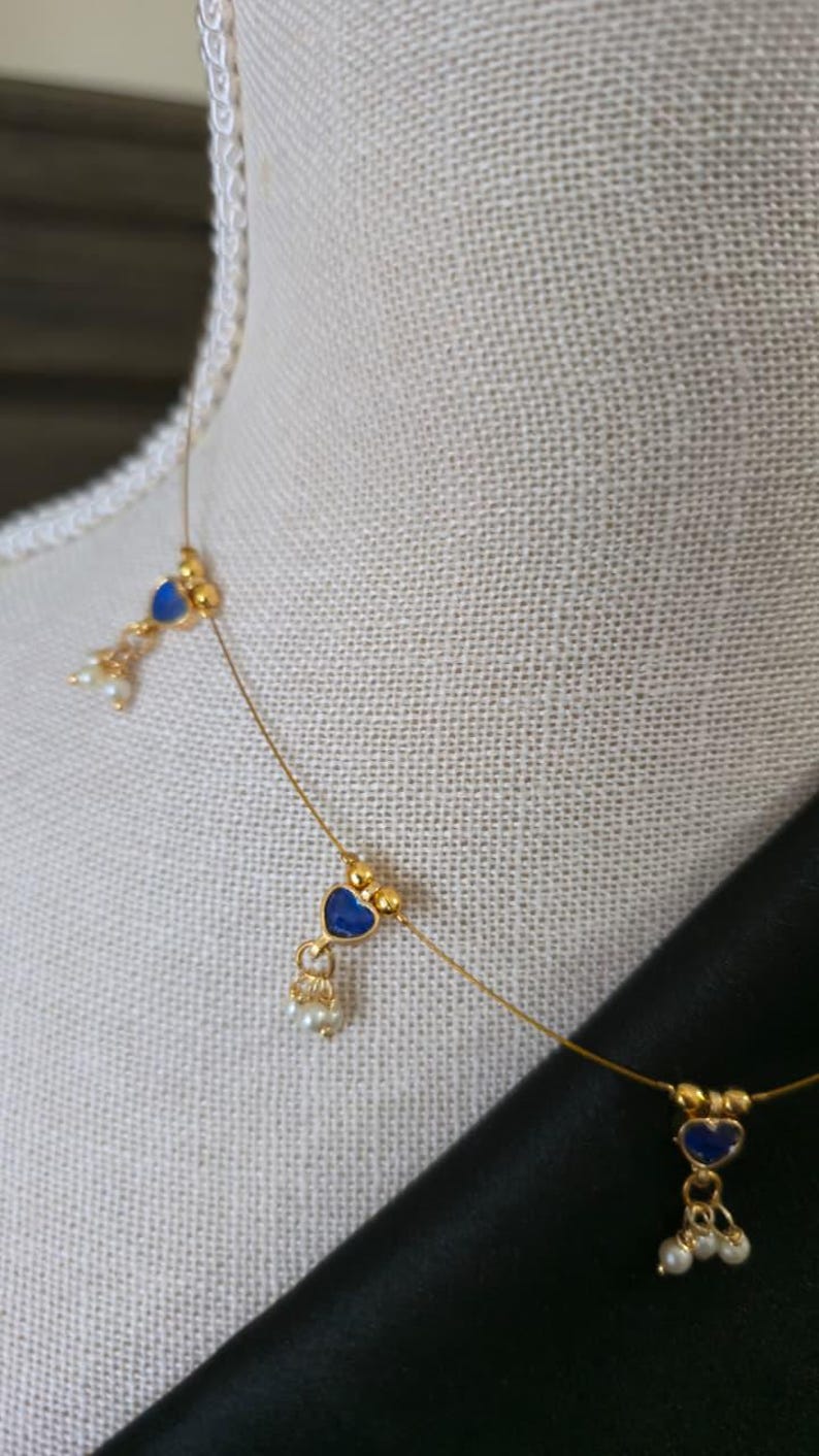 Puede incluir: Delicado collar de oro con colgantes azules en forma de coraz&oacute;n y peque&ntilde;os detalles de perlas blancas. El collar se exhibe en un maniqu&iacute;, mostrando su elegante dise&ntilde;o. Una joya elegante.