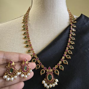 May include: A gold-toned necklace and earring set. The necklace features a series of green and red gemstones, with a central pendant of a larger green stone and white pearls. The earrings mirror the pendant design, with a cluster of white pearls.