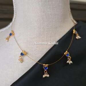 Puede incluir: Un delicado collar dorado con colgantes azules en forma de coraz&oacute;n y peque&ntilde;os detalles de perlas blancas. El collar se exhibe en un maniqu&iacute;, con el texto "Namma Kadai" visible.