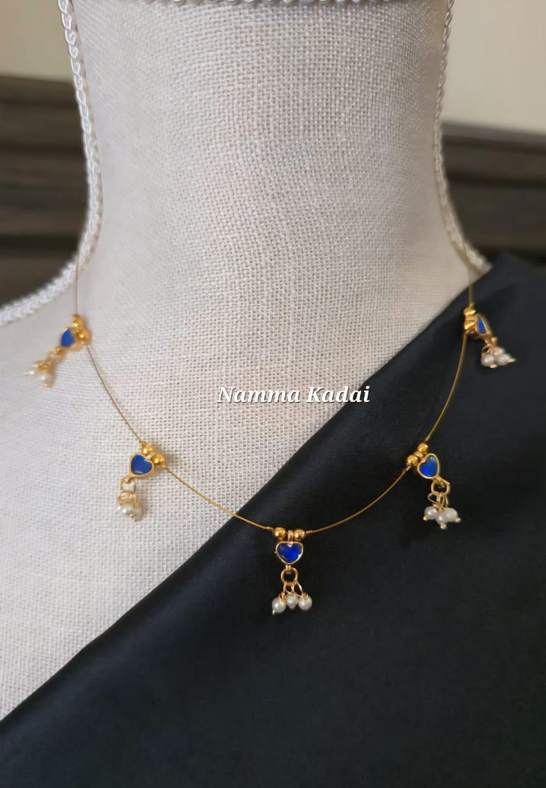 Puede incluir: Un delicado collar dorado con colgantes azules en forma de coraz&oacute;n y peque&ntilde;os detalles de perlas blancas. El collar se exhibe en un maniqu&iacute;, con el texto "Namma Kadai" visible.