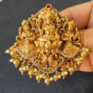May include: A gold-colored pendant featuring three figures, with intricate detailing and small white pearls hanging from the bottom. The pendant has a central figure seated on a lotus flower, flanked by two standing figures.