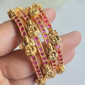 May include: A set of four gold-toned bangles featuring a series of square-cut pink stones embedded along the band. Each bangle is accented with a raised, detailed gold-toned figure of a deity, providing a textured and ornate appearance to the jewelry.