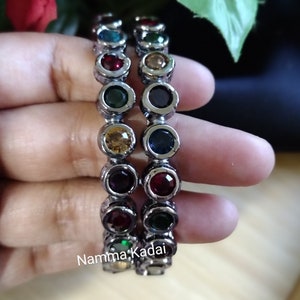 May include: Two silver bangles with colorful round stones. The stones are arranged in a rainbow pattern.