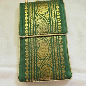 May include: A rectangular green fabric pouch featuring gold metallic woven patterns of paisley and floral motifs. The pouch has a fold-over flap, gold piping trim along the edges, and a thin gold-toned metal chain strap attached to a metal clasp.