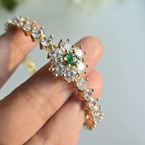 Puede incluir: Una pulsera de tono dorado con un adorno central en forma de flor compuesto por piedras transparentes de corte redondo que rodean una peque&ntilde;a piedra redonda de color verde vibrante. La banda presenta una hilera continua de piedras claras.