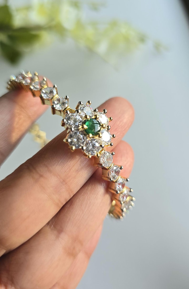 Puede incluir: Una pulsera de tono dorado con un adorno central en forma de flor compuesto por piedras transparentes de corte redondo que rodean una peque&ntilde;a piedra redonda de color verde vibrante. La banda presenta una hilera continua de piedras claras.