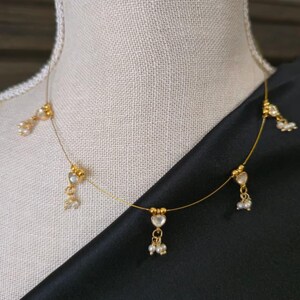 Puede incluir: Delicado collar dorado con colgantes en forma de coraz&oacute;n y peque&ntilde;os detalles de perlas. El collar se exhibe sobre un maniqu&iacute;, con un fondo de tela negra. El collar tiene un dise&ntilde;o minimalista, con una cadena fina y m&uacute;ltiples elementos decorativos.